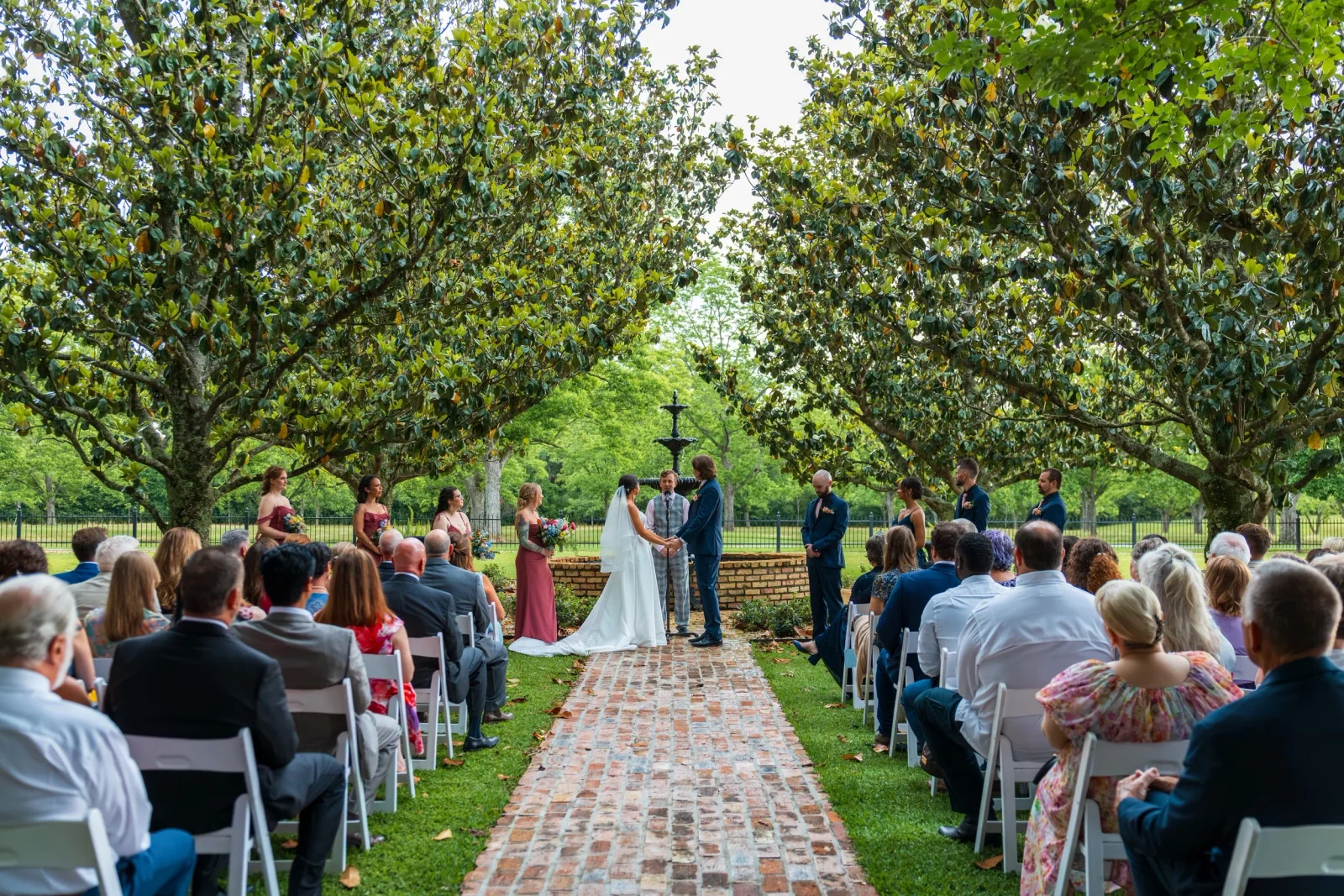 A couple exchange vows at Magnolia Fountain, with guests seated beneath the trees.